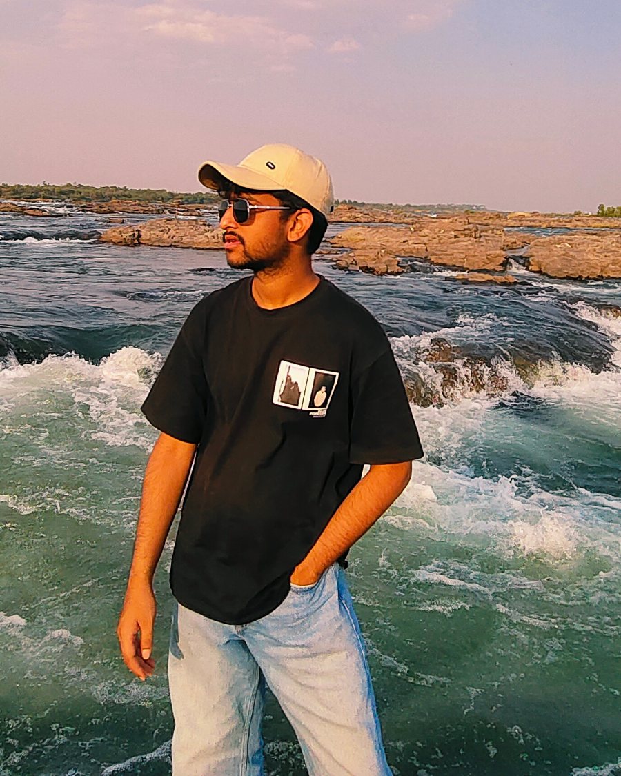 Parth Tiwari standing in front of river rapids at golden hour, in a black tee, pale jeans, sunglasses and a cream cap, looking off to one side.